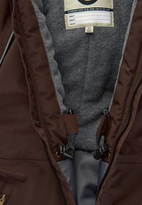 Chaqueta exterior marrón con un forro de forro polar gris, que presenta cierres de cremallera, toggles de cordón y una etiqueta con las instrucciones de cuidado.