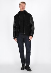 Veste noire en daim et cuir, avec fermeture éclair à l'avant, col en shearling et manches en cuir côtelé, associée à un jean foncé et des bottes.