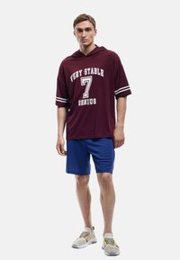 Maroon capuchonshirt met witte tekst en strepen, gecombineerd met blauwe shorts. Schoenen zijn grijs met gele accenten.