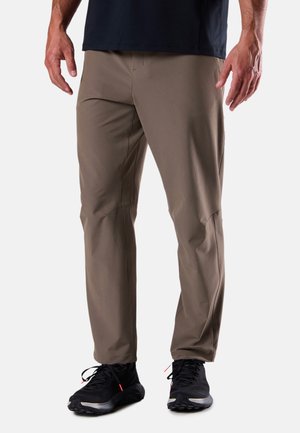 Mann trägt dunkelbraune, schlanke Hose und schwarze Sportschuhe, steht vor einem schlichten hellen Hintergrund.