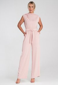 Różowy kombinezon z wysokim dekoltem, bez rękawów, elastyczną talią i paskiem do zawiązania. Posiada szerokie nogawki i boczne kieszenie. Gładka struktura materiału.