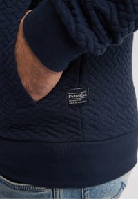 Mörkblå quiltad sweatshirt med ribbad mudd och nederkant. Har en sidoficka och en märkesetikett sydd i tyget. Mjuk textur.