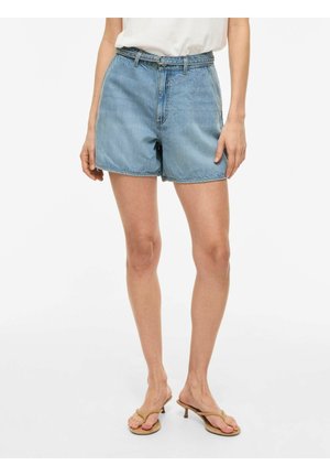 Kvinne iført lyseblå høytlivs denimshorts med tynn belte, hvit topp og beige sandaler med hæl, står foran en ensfarget bakgrunn.