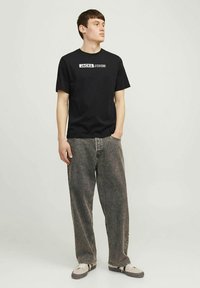 Juodas medvilninis marškinėlis su priekyje baltu logotipu „JACK & JONES“, derinamas su tamsiai pilkais plačiomis kojomis džinsais ir šviesios spalvos bateliais.