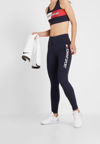 Marina leggings med 'TOMMY SPORT' tryckt i vitt, tillsammans med en matchande sportbh med ett rött rand. Modellen håller en vit jacka.