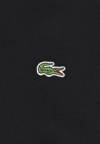 Logo de crocodile vert brodé avec un détail de bouche rouge sur un fond de tissu côtelé noir centré dans le cadre.