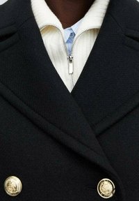 Abrigo negro cruzado con botones dorados y solapas anchas sobre un suéter blanco de cremallera y canalé, con cuello de camisa azul claro.