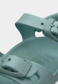 Τυρκουάζ σανδάλι με χοντρές, ευλύγιστες λουρίδες και ρυθμιζόμενο μεταλλικό πόρπη. Μαλακή, υφή επιφάνεια· το διαμορφωμένο πέλμα φέρει αποτύπωμα λογότυπου.