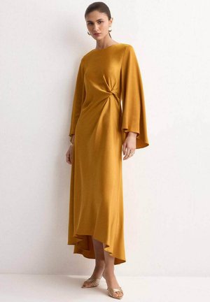 Mujer modelando un vestido de manga larga de color mostaza con un detalle de nudo lateral y dobladillo asimétrico, llevando pendientes dorados y tacones abiertos.