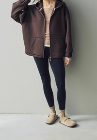 Femme portant un sweat à capuche marron surdimensionné avec fermeture éclair, haut beige, leggings noirs, chaussettes beiges et chaussures beiges à enfiler, debout contre un mur gris.