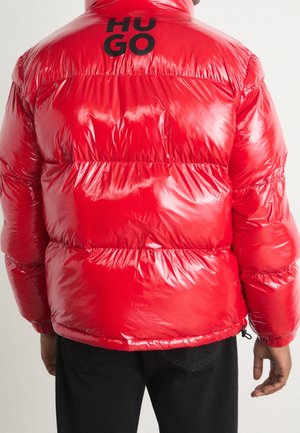 Personne portant une doudoune rouge brillant avec le logo noir "HUGO" sur le haut du dos, associée à un pantalon noir, tournant le dos à la caméra.