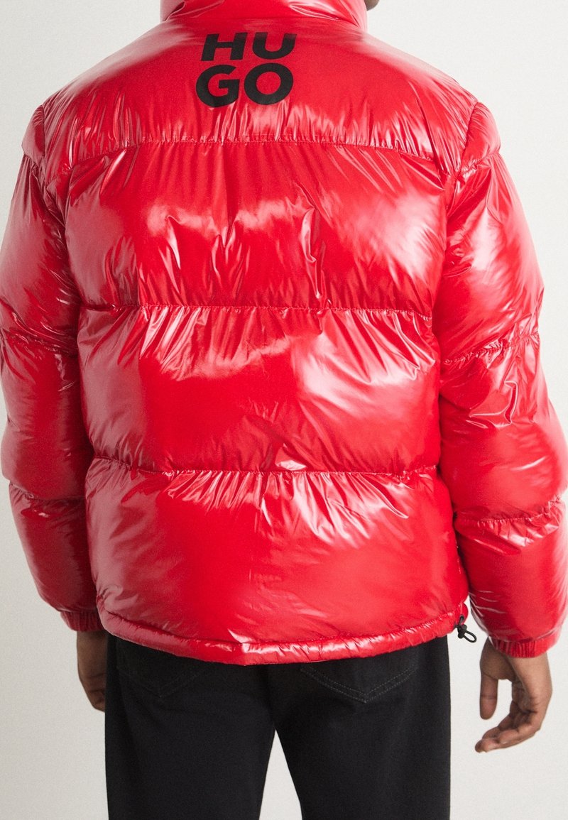 Personne portant une doudoune rouge brillant avec le logo noir "HUGO" sur le haut du dos, associée à un pantalon noir, tournant le dos à la caméra.
