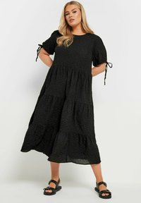 Svart lager-på-lager klänning i texturerad tyg, med korta ärmar som har knytningar. Modellen bär svarta sandaler. Minimalistisk design utan mönster.