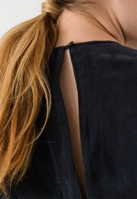 Černý textilní oděv s výřezem ve tvaru klíče na zádech, ukazující hladkou texturu a měkký splývavý střih, kontrastující s vlnícími se vlasy.