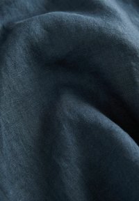 Tmavozelená ľanová tkanina s jemným, textúrovaným povrchom, s miernymi prehybmi vytvárajúcimi hĺbku a jemné variácie v odtieňoch.