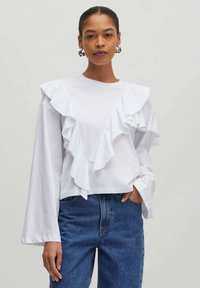 Weißes Langarmoberteil mit gestuften Rüscheln an der Vorderseite. Kombiniert mit blauen Jeans aus Denim, die einen lässigen, strukturierten Schnitt zeigen.