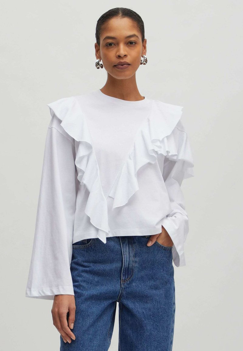 Weißes Langarmoberteil mit gestuften Rüscheln an der Vorderseite. Kombiniert mit blauen Jeans aus Denim, die einen lässigen, strukturierten Schnitt zeigen.