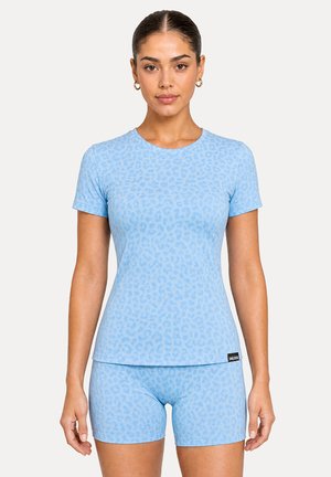 Femme portant un t-shirt ajusté et un short assortis à imprimé léopard bleu clair, debout devant un fond clair uni.