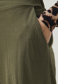 Ελαιοπράσινο ύφασμα από παντελόνι σε άνετη γραμμή με ορατή πλευρική τσέπη. Η υφή φαίνεται μαλακή και ελαφρώς ζαρωμένη, με χαλαρό σχέδιο.