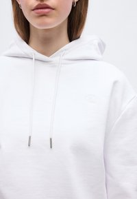 Jeune personne portant un sweat à capuche blanc avec cordons et petit logo "cK" sur la poitrine, montrant la partie inférieure du visage et le cou avec des boucles d'oreilles anneaux.