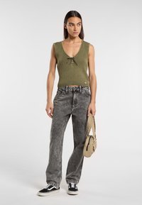 Top de tirantes de color verde oliva con detalle de nudo en la parte delantera, combinado con jeans de tiro alto en lavados ácidos grises y zapatillas negras, sosteniendo un pequeño bolso beige.