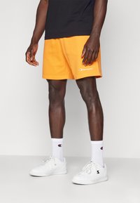 Oranžid puuvillased lühikesed püksid paelaga vööl ja broderitud "Champion" logo. Kandmine valgete sokkide ja valgete spordijalatsitega, seistes heledal taustal.