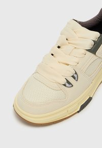 Zapatilla beige con malla texturizada y gamuza suave, cordones anchos color crema y un sutil acento oscuro cerca de los ojetes. Suela de goma.