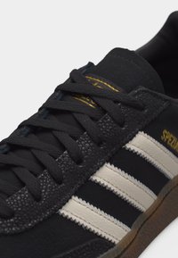 Μαύρο αθλητικό παπούτσι Adidas Spezial με λευκές ζιγκ ζαγκ ρίγες, μαύρα κορδόνια, λεπτομέρεια από δέρμα με υφή και καφέ σόλα από καουτσούκ.
