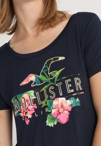 Mörkblå skjorta med blommönstrad Hollister California-logotyp, bärs av en person med ljus hy och axellångt blont hår.