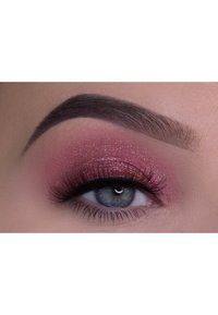 Close-up van een oog met roze glinsterende oogschaduw, lange zwarte wimpers, goed gedefinieerde donkere wenkbrauw en blauwig-groene iris.