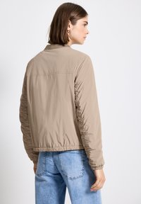Veste beige légère avec des poignets élastiques et un col côtelé, présentant des détails froncés sur les manches. Portée avec un jean en denim bleu clair.