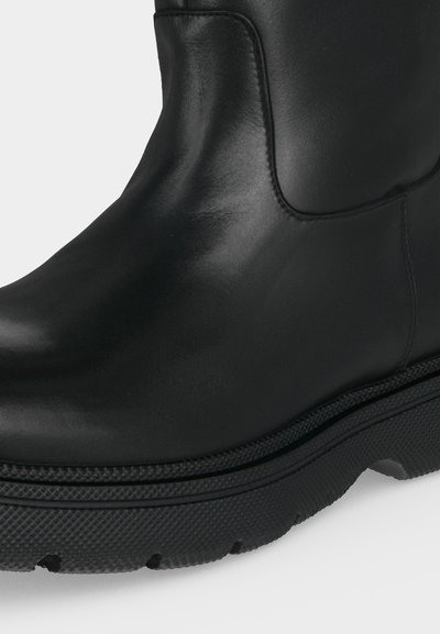 Bottine en cuir noir avec semelle épaisse en caoutchouc texturée et dessus lisse avec coutures visibles sur un fond uni.