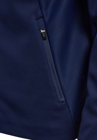 Close-up van een zijzak met rits aan een marineblauwe jas, met een stoffen trekkoord voorzien van twee chevron-pijlen.