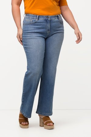 Person iført medium-blå jeans, brune åben tå kile-sandaler og en orange top, der står mod en ensartet lys baggrund.