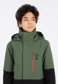 Giacca verde leggera con accenti neri, colletto alto, tasca sul petto con zip e dettagli del logo. Presenta un cappuccio regolabile e zip pull.