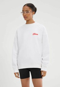 Mujer con gafas transparentes, sudadera blanca con el logo rojo de "Ellesse" y pantalones cortos negros, de pie frente a un fondo claro y liso.