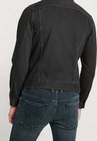 Giacca di jeans nera con un design leggermente strutturato, caratterizzata da una spalla posteriore e dettagli sulle cuciture laterali, abbinata a jeans blu scuro.