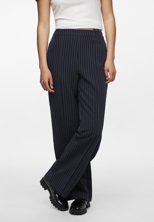 Vrouw in zwarte pinstripe wijde broek met zwarte grove schoenen en een wit cropped topje, staand met gekruiste benen.