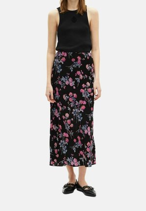 Femme portant un haut noir sans manches, une longue jupe noire avec un motif floral rose et bleu, et des chaussures plates noires.