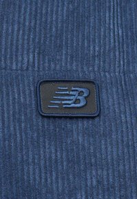 Modrý kordurojový materiál s vertikálnym ribovaním; obsahuje čiernu nášivku loga New Balance s modrým prešívaním. Mäkkučká textúra, bez ďalších vzorov.