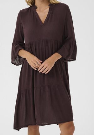 Femme portant une robe ample marron foncé à volants, manches cloche trois-quarts et petit décolleté en V avec encoche, posant devant un fond uni.