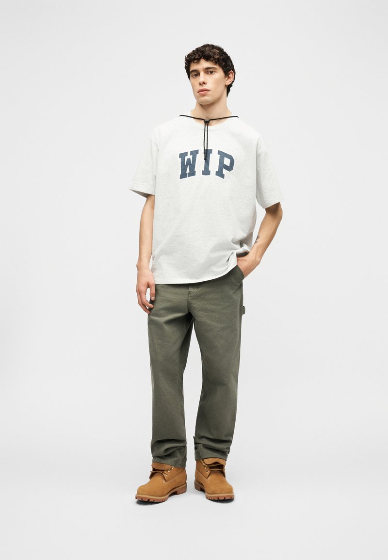 Junger Mann steht mit einer Hand in der Tasche, trägt ein weißes WIP-T-Shirt, olivgrüne Hosen und beige Arbeitsstiefel vor einem einfarbigen Hintergrund.