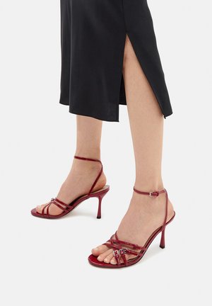 Czerwone sandały na wysokim obcasie z lakierowanej skóry charakteryzują się cienkimi paskami, klamrą przy kostce oraz spiczastym noskiem. Obcas ma około 8 cm wysokości.