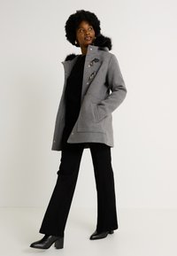 Abrigo de lana gris con capucha forrada de piel, detalles en hardware dorado, grandes bolsillos frontales y un corte entallado, combinado con pantalones negros acampanados y tacones.