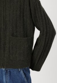 Dark green knitted cardigan with ribbed texture, front zipper, and a single patch pocket on the left side. Worn over blue denim.