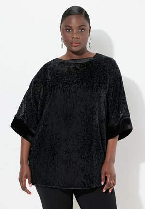 Top de terciopelo negro con un patrón floral en relieve, mangas anchas y un escote redondo. Combinado con pantalones negros y grandes pendientes de aro.