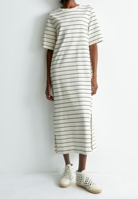 Osoba ve smetanových midi šatech s černými horizontálními pruhy a bočními rozparky, doplněná bílými pletenými kotníkovými botami se šněrováním.