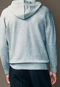 Lichtgrijze hoodie met een zachte textuur, voorzien van een trekkoordcapuchon en ribbels aan de onderkant. Gecombineerd met donkere pantalon met streepjes.