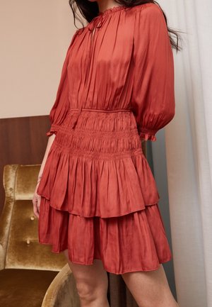 Kobieta w długiej, rudobrązowej sukience z falbanami, stojąca obok złotego aksamitnego krzesła i białej zasłony.