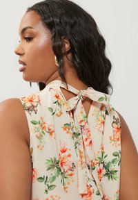 Blusa floreale con sfondo crema, caratterizzata da fiori arancioni e foglie verdi. Include un dettaglio con legaccio sul retro e ha un design senza maniche.